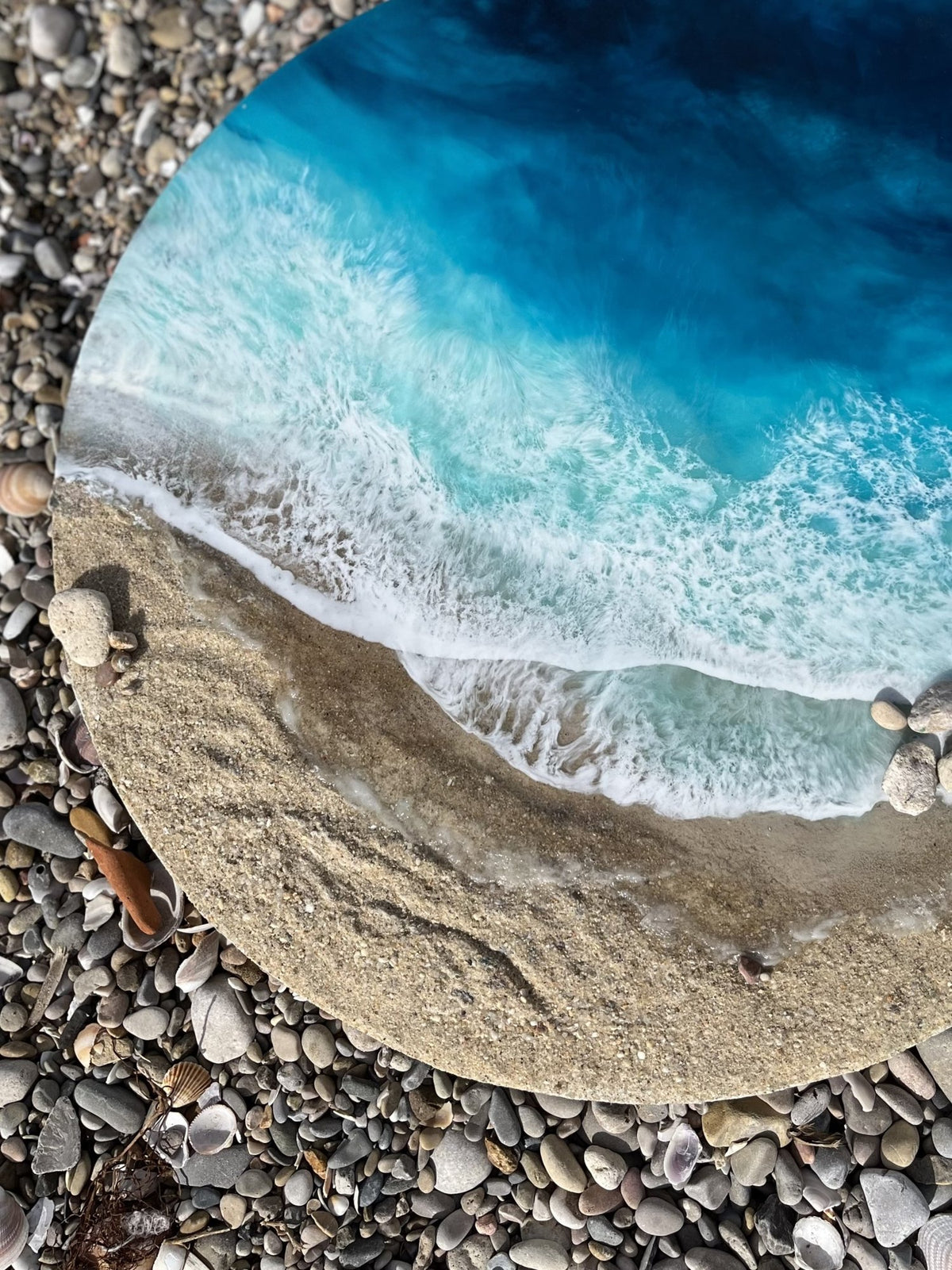 Obra redonda de Ocean Art 3D de Eva Jurado sobre guijarros: mar turquesa, espuma blanca y orilla de arena